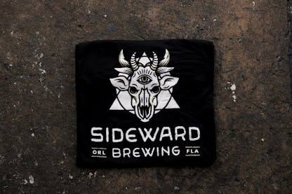 A black tee featuring a white Sideward Brewing logo, which resembles a demon cow skull with a triangle and circle behind it, with the Sideward Brewing wordmark below.