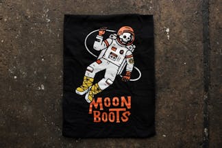 A black tee with a skeleton astronaut with the words 'Moon Boots' underneath in burnt orange.