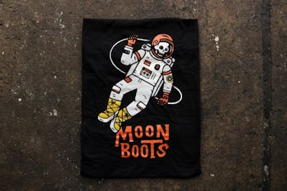 A black tee with a skeleton astronaut with the words 'Moon Boots' underneath in burnt orange.