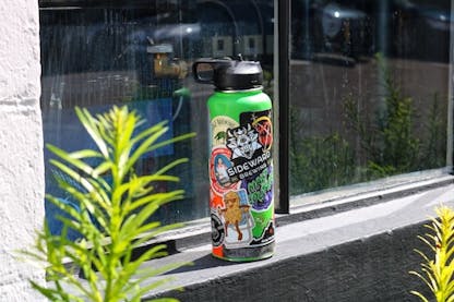 A green water bottle covered in various stickers featuring a prominent Sideward Brewing logo sticker.