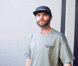 Man wearing a hat and a sage shirt with the words, Slow Pours Big Foam