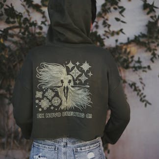 Women wearing cropped olive green hooded sweatshirt with intricate design in cream. Design is two women back to back with long hair waving in the wind and decorative stars around them. Below them it says "ex novo brewing company"