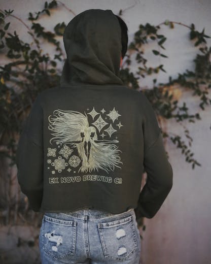 Women wearing cropped olive green hooded sweatshirt with intricate design in cream. Design is two women back to back with long hair waving in the wind and decorative stars around them. Below them it says "ex novo brewing company"