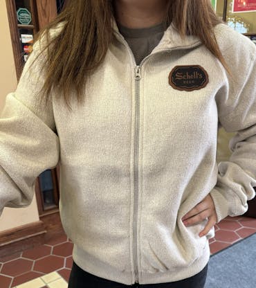 Oatmeal colored full zip sweater with Schell logo on front left lapel.