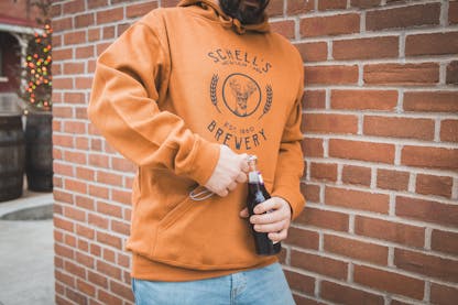 Copper colored sweatshirt with Schell logo on front and a bottle coozie in front pocket.