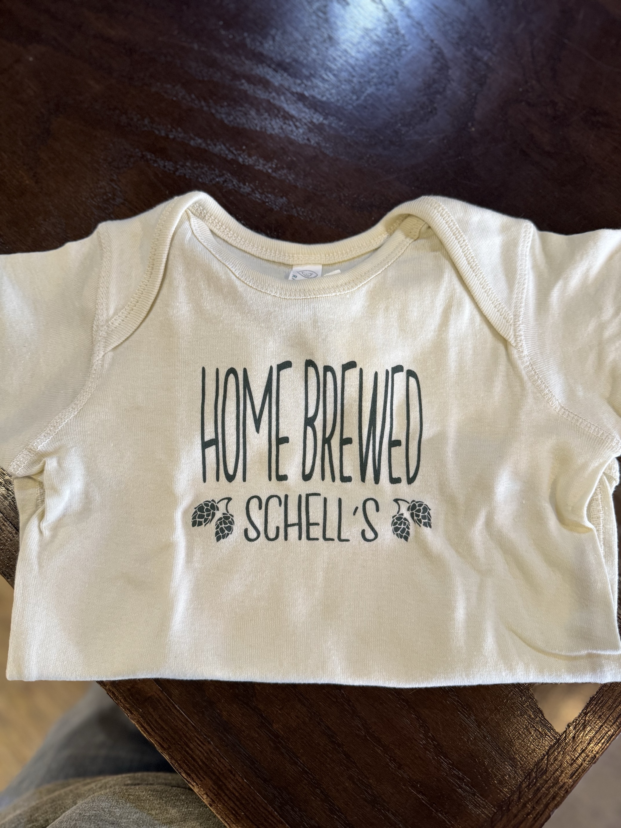 Cream colored infant onesie with Schell logo on front.