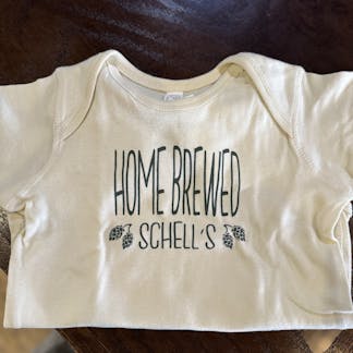 Cream colored infant onesie with Schell logo on front.