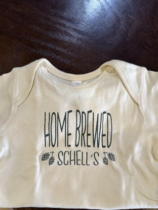 Cream colored infant onesie with Schell logo on front.