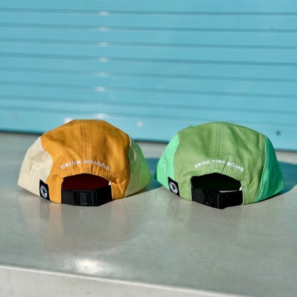the back of a blue and green hat that reads "drink Tiny Bomb" next to the back of red and orange hat that says "drink Ananda"