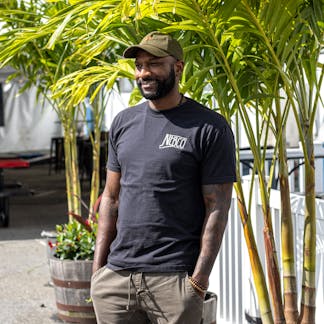 male model wearing black t-shirt in front of palm tree with NEBCO state logo on chest