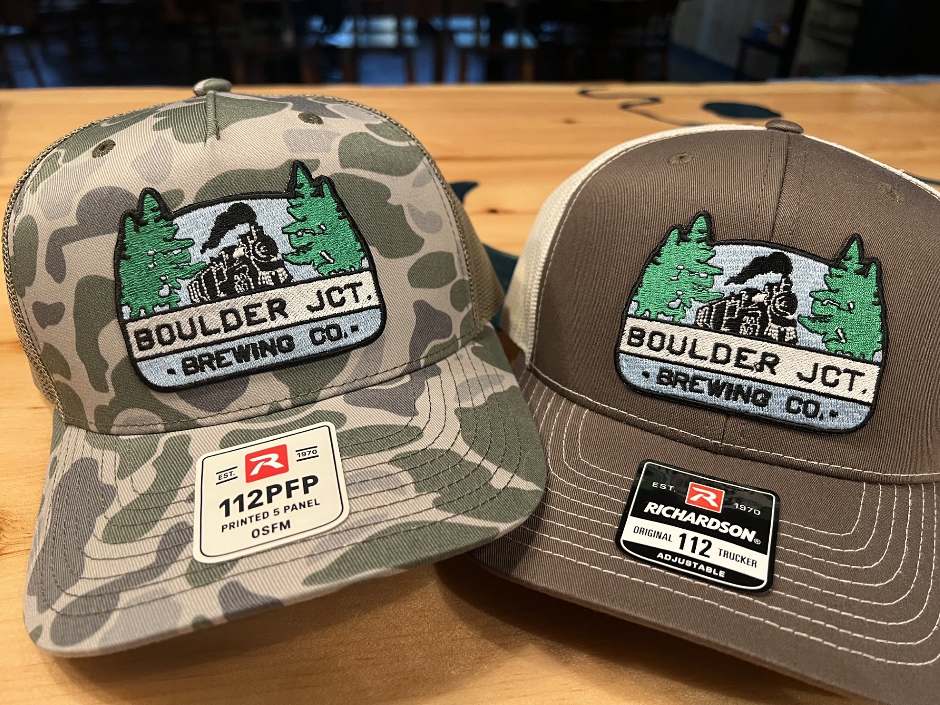 2 trucker hats on the bar, one is chocolate the other is camo and both have embroidered logo patches on the front