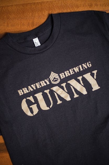 front detail of Gunny black t-shirt flat on a wooden table. The text "Bravery Brewing Gunny" along with icon art from the Gunny beer can is printed large across the chest in light brown ink.