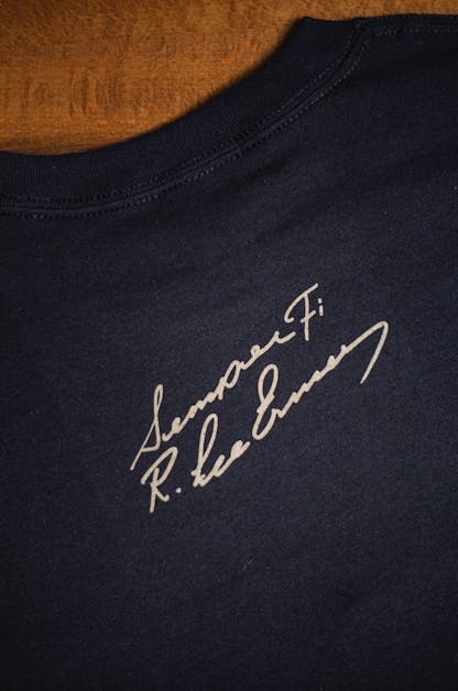 back detail of Gunny black t-shirt flat on a wooden table. The text "Semper Fi R. Lee Ermey" in a handwritten script is printed small below the back collar in light brown ink.