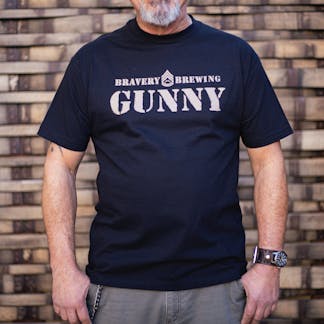 front of Gunny black t-shirt worn by a male model. The text "Bravery Brewing Gunny" along with icon art from the Gunny beer can is printed large across the chest in light brown ink.