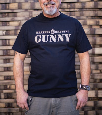 front of Gunny black t-shirt worn by a male model. The text "Bravery Brewing Gunny" along with icon art from the Gunny beer can is printed large across the chest in light brown ink.