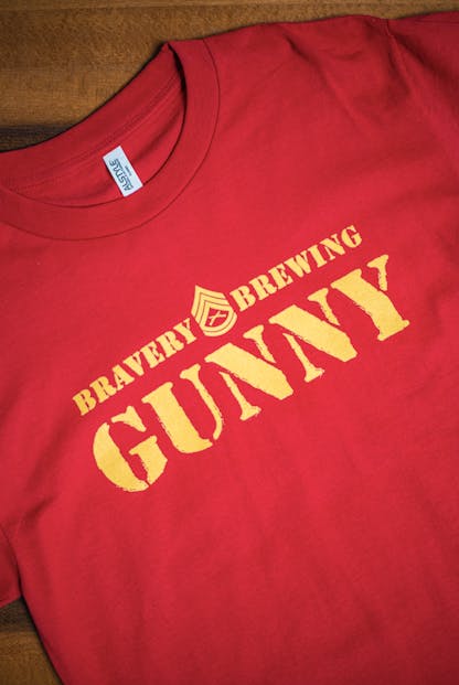 front detail of Gunny red t-shirt flat on a wooden table. The text "Bravery Brewing Gunny" along with icon art from the Gunny beer can is printed large across the chest in yellow ink.