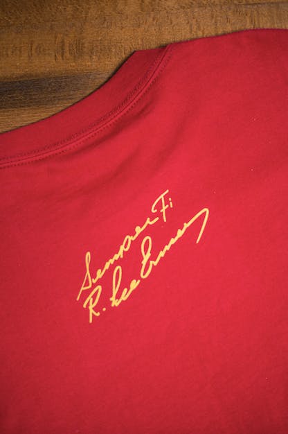 back detail of Gunny red t-shirt flat on a wooden table. The text "Semper Fi R. Lee Ermey" in a handwritten script is printed small below the back collar in yellow ink.
