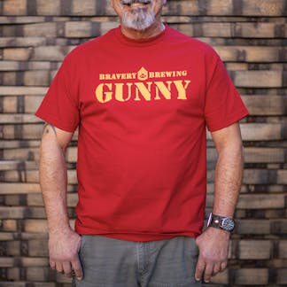 front of Gunny red t-shirt worn by a male model. The text "Bravery Brewing Gunny" along with icon art from the Gunny beer can is printed large across the chest in yellow ink.