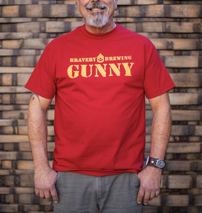 front of Gunny red t-shirt worn by a male model. The text "Bravery Brewing Gunny" along with icon art from the Gunny beer can is printed large across the chest in yellow ink.