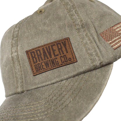 Detail photo of the front side of a green baseball cap on a white background. A brown leather suede patch with the text "Bravery Brewing Co." is embroidered on the front left panel of the hat. An American flag is embroidered on the side panel with brown thread.