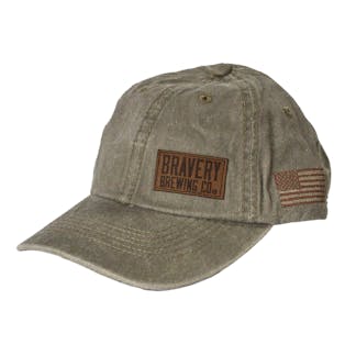 Front side photo of light green baseball cap on a white background. The hat is a worn weathered material with a brown embroidered flag on the side, and a leather suede patch sewn on to the front left panel of the hat.