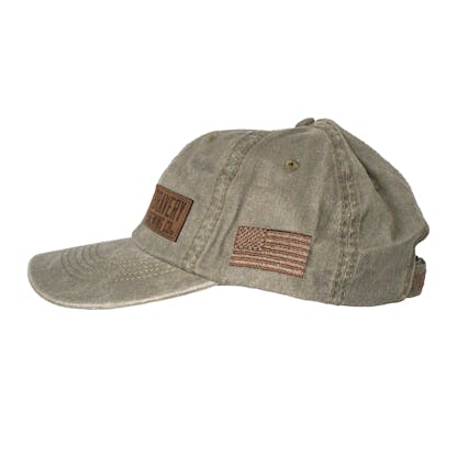 Side photo of light green baseball cap on a white background. The hat is a worn weathered material with a brown embroidered flag on the side, and a leather suede patch sewn on to the front left panel of the hat.
