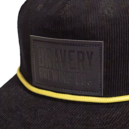 Front detail photo of a black corduroy snapback on a white background. The hat has a thick yellow cord along where the brim meets the hat, and a black leather patch on the front two panels. The leather patch has the logo for Bravery etched on it.