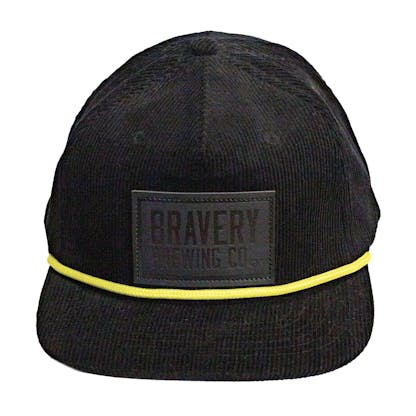 Front photo of a black corduroy snapback on a white background. The hat has a thick yellow cord along where the brim meets the hat, and a black leather patch on the front two panels. The leather patch has the logo for Bravery etched on it.
