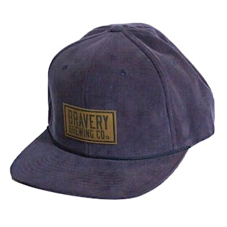 Front side photo of a Blue corduroy snapback on a white background. The hat has a thick blue cord along where the brim meets the hat, and a brown leather patch on the front two panels. The leather patch has the logo for Bravery etched on it.