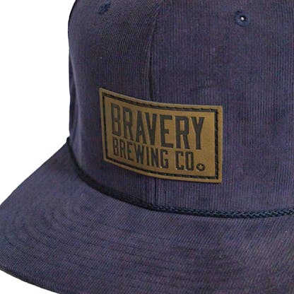 Front detail photo of a blue corduroy snapback on a white background. The hat has a thick blue cord along where the brim meets the hat, and a brown leather patch on the front two panels. The leather patch has the logo for Bravery etched on it.