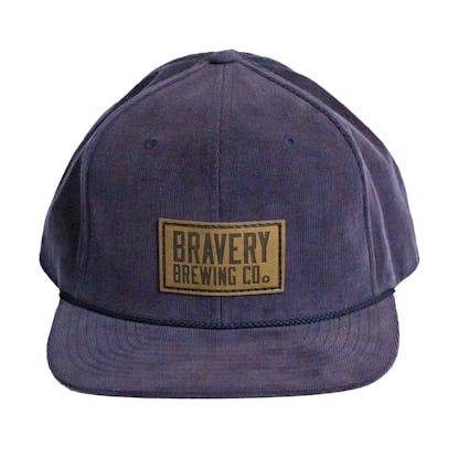 Front photo of a blue corduroy snapback on a white background. The hat has a thick blue cord along where the brim meets the hat, and a brown leather patch on the front two panels. The leather patch has the logo for Bravery etched on it.