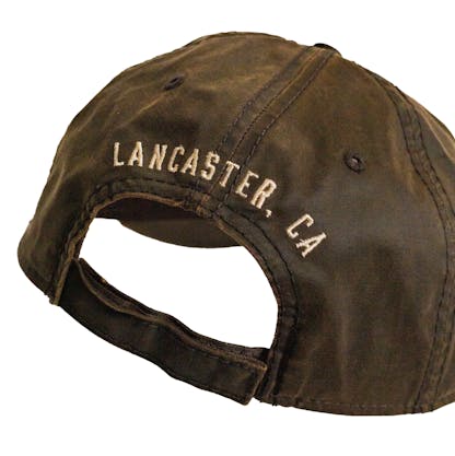 Back detail photo of brown baseball cap on a white background. The hat is a lightly worn weathered material with white embroidered text across the back two panels that says "LANCASTER, CA"