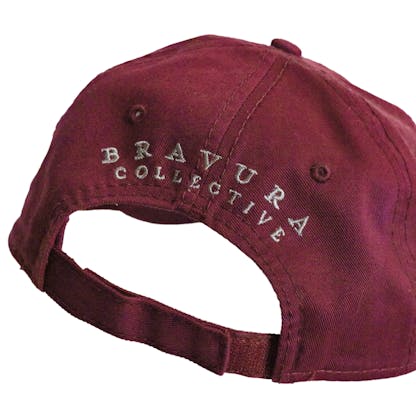 Back detail photo of maroon baseball cap on a white background. The fabric hat has grey embroidered text across the back two panels that says "BRAVURA COLLECTIVE"