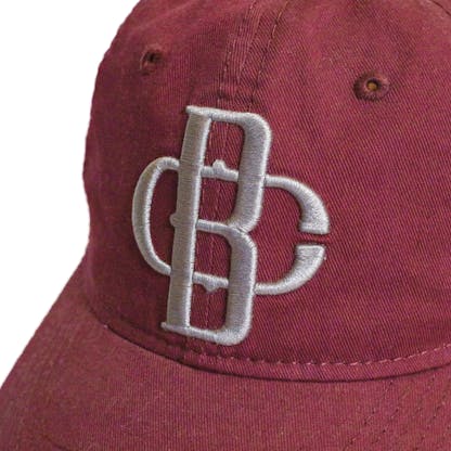Front detail photo of maroon baseball cap on a white background. The fabric hat has a grey embroidered logo for the Bravura Collective centered on the front two panels.