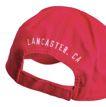 Back detail photo of bright pink baseball cap on a white background. The fabric hat has white embroidered text across the back two panels that says "LANCASTER, CA"