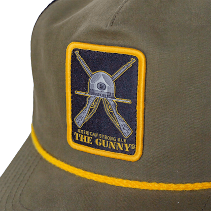 Detail photo of green trucker cap on a white background. The hat has green fabric for the front two panels. There is a white, grey and gold embroidered patch with art for the beer "The Gunny" across the front two panels. There is a yellow cord on the edge of the hat where the bill meets the cap.