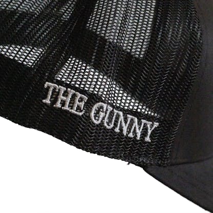 Side detail photo of Black trucker cap on a white background. The right side mesh has the text "THE GUNNY" embroidered in white along the bottom edge of the hat.