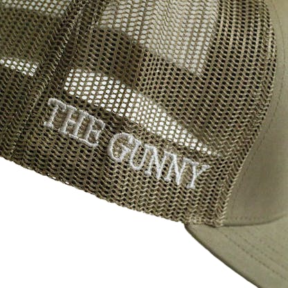 Side detail photo of green trucker cap on a white background. The right side mesh has the text "THE GUNNY" embroidered in white along the bottom edge of the hat.