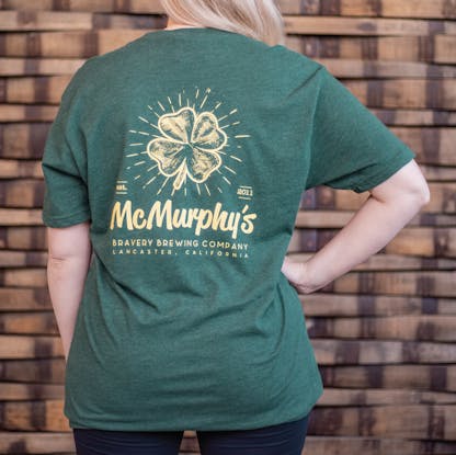 Photo of the back of a person wearing a t-shirt. The t-shirt is a deep heather green with an illustration and text printed in light yellow on the back. The art is of a four leaf clover with the text "McMurphy's Bravery Brewing Company Lancaster California, est. 2011" underneath it.