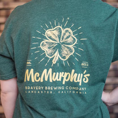 Detail photo of the back of a person wearing a t-shirt. The t-shirt is a deep heather green with an illustration and text printed in light yellow on the back. The art is of a four leaf clover with the text "McMurphy's Bravery Brewing Company Lancaster California, est. 2011" underneath it.