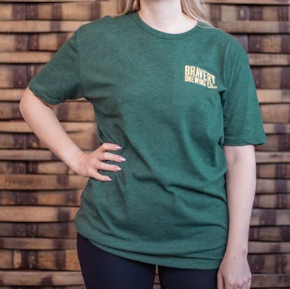 Photo of the front of a person wearing a t-shirt. The t-shirt is a deep heather green with text printed in light yellow on the wearers left chest. The text says "Bravery Brewing Company"