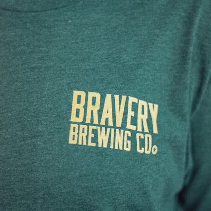 Detail photo of the front of a person wearing a t-shirt. The t-shirt is a deep heather green with text printed in light yellow on the wearers left chest. The text says "Bravery Brewing Company"