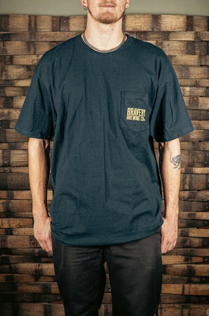 Photo of a model wearing a navy t-shirt. The t-shirt has a pocket on the wearer's left side. On the pocket there is the text "Bravery Brewing Company" printed in yellow ink.
