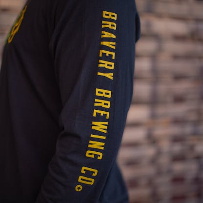 Detail photo of the sleeve of a person wearing a black long sleeve. The sleeve has the text "Bravery Brewing Co." printed down the sleeve in yellow ink.