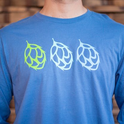 Detail photo of a person wearing a light blue long sleeve shirt from the front. The front of the shirt has the icons of three duplicate hops across the chest, from left to right they are green, white, and pale blue in color.