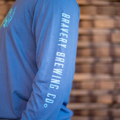 Detail photo of the sleeve of a person wearing a light blue long sleeve. The sleeve has the text "Bravery Brewing Co." printed down the sleeve in pale blue ink.