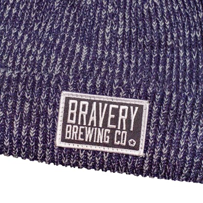 Detail photo of a knit beanie on a white background with a black and white embroidered patch sewn on the front. The hat is a blue and white marbled yarn and the patch has the text "Bravery Brewing Co." embroidered in white onto it.