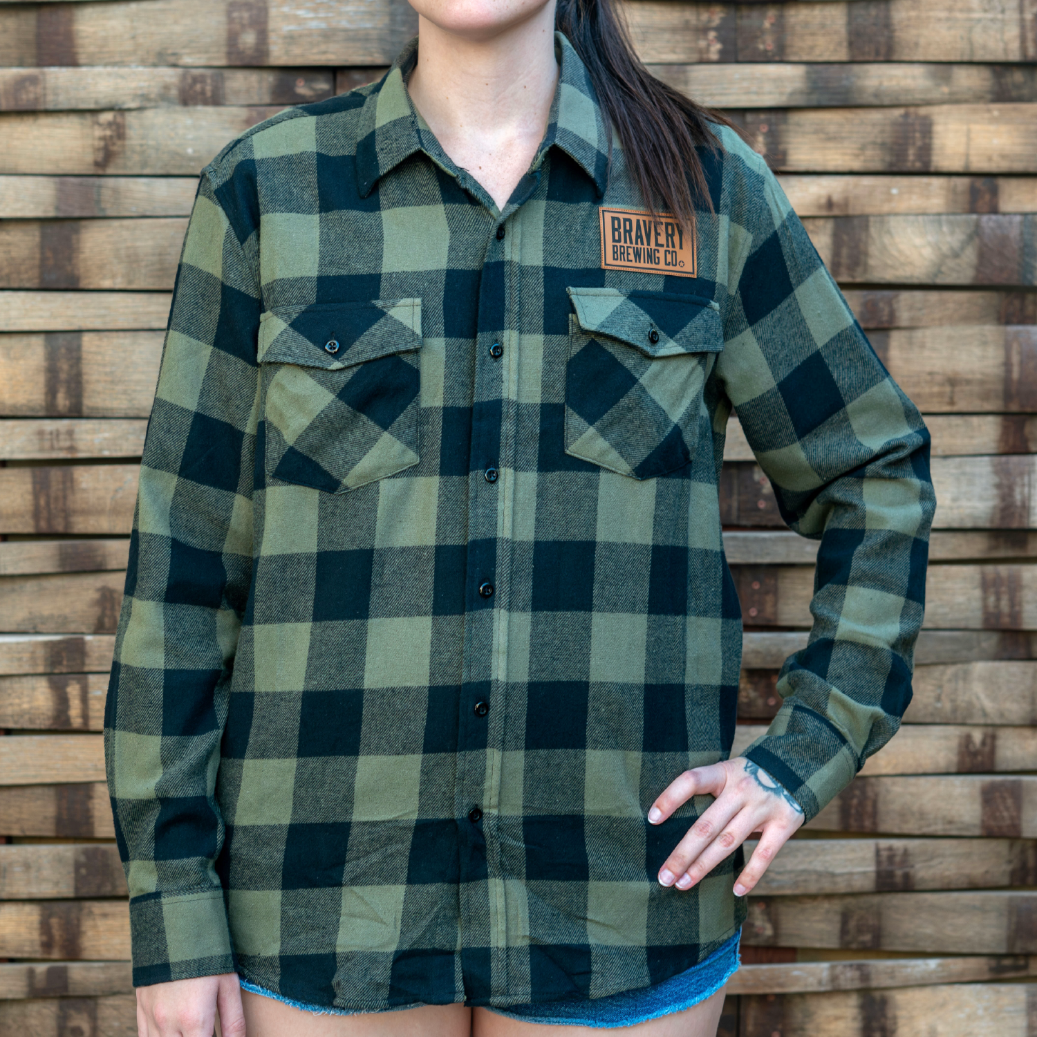 front of green and black Flannel shirt worn by a model. Above the wearer's left chest pocket is an leather patch with Bravery Brewing Co.'s logo.