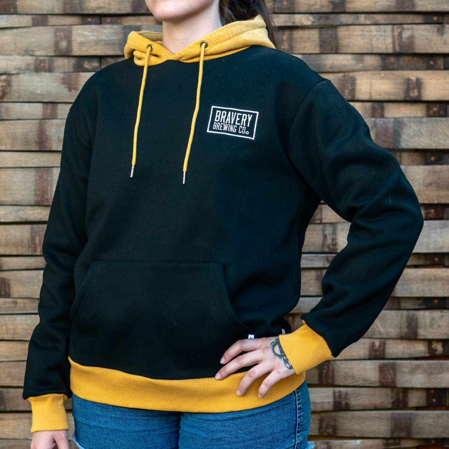 Front of black and gold hoodie worn by a model. Above the wearer's left chest pocket is Bravery Brewing Co.'s logo embroidered in white. The hood and cuffs are gold while the body and sleeves are black.